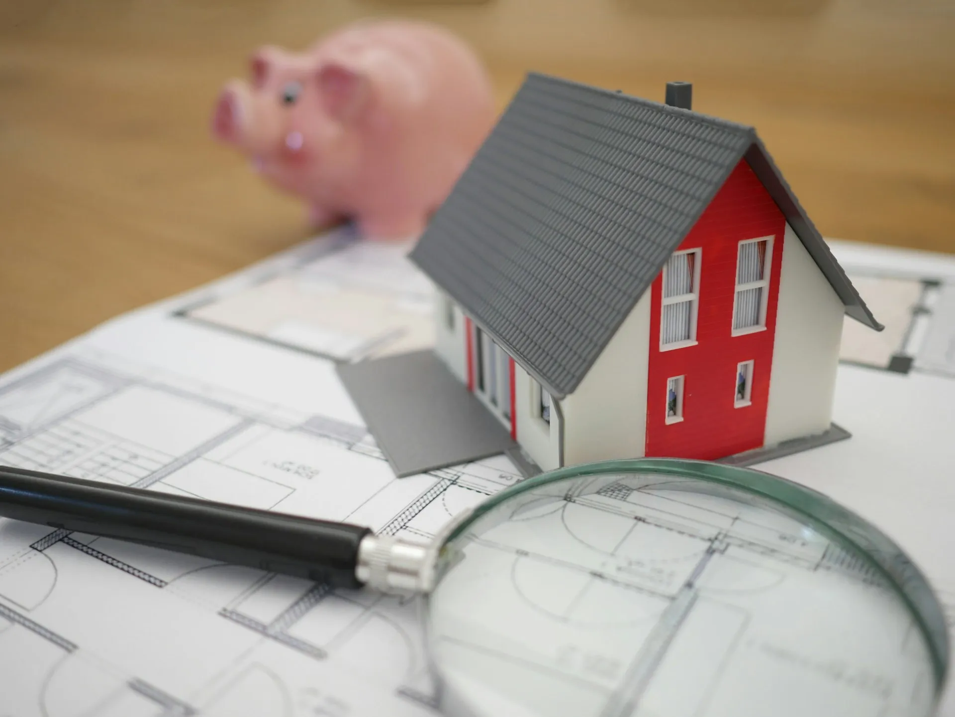 Modellhaus mit rotem Giebel auf Bauplänen, daneben Lupe und Sparschwein