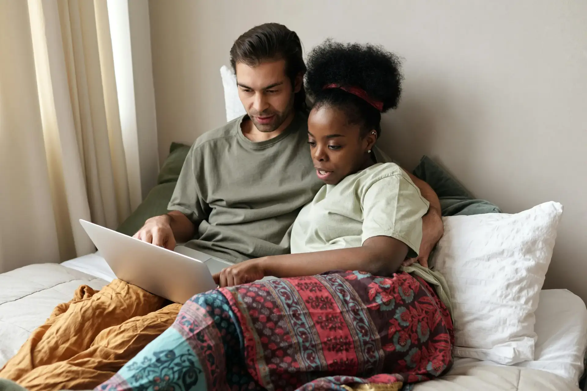 Ein Paar sitzt gemütlich zusammen auf dem Bett und arbeitet gemeinsam an einem Laptop, möglicherweise um Informationen zur Baufinanzierung und Hausfinanzierung zu recherchieren, was Nähe und Verbundenheit in der modernen, digitalen Welt zeigt.